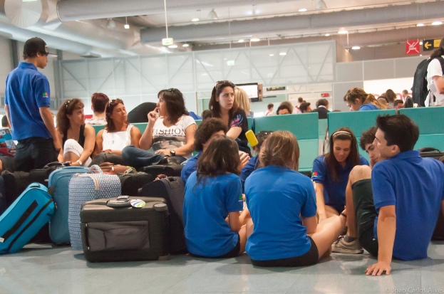 Ein belebter Flughafen-Terminal mit vielen Menschen, die auf ihre Flüge warten, Gepäckstücke links daneben und Richtungsschilder sowie Glastüren im Hintergrund.