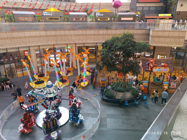 Ein Vergnügungspark innerhalb eines Einkaufszentrums mit verschiedenen Attraktionen, Bäumen, Menschen und Ständen, umgeben von Gebäuden, mit einem Wasserzeichen in der unteren rechten Ecke.