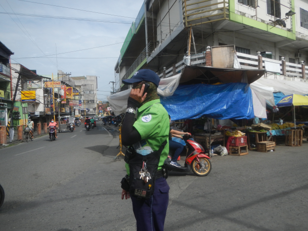 Ein Polizeibeamter in grüner und blauer Uniform und blauer Mütze steht an der Straße, spricht in ein Handy, mit Fahrzeugen, die hinter ihm fahren, Ständen mit verschiedenen Gegenständen zu seiner Rechten und einem bewölkten Himmel mit einem Gebäude im Hintergrund.