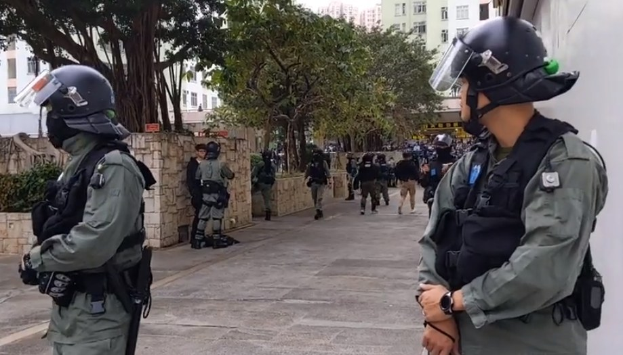 Eine Gruppe von Polizisten in Helmen und kugelsicheren Westen steht auf einer Straße in Hong Kong, mit Bäumen, Gebäuden und dem Himmel im Hintergrund.