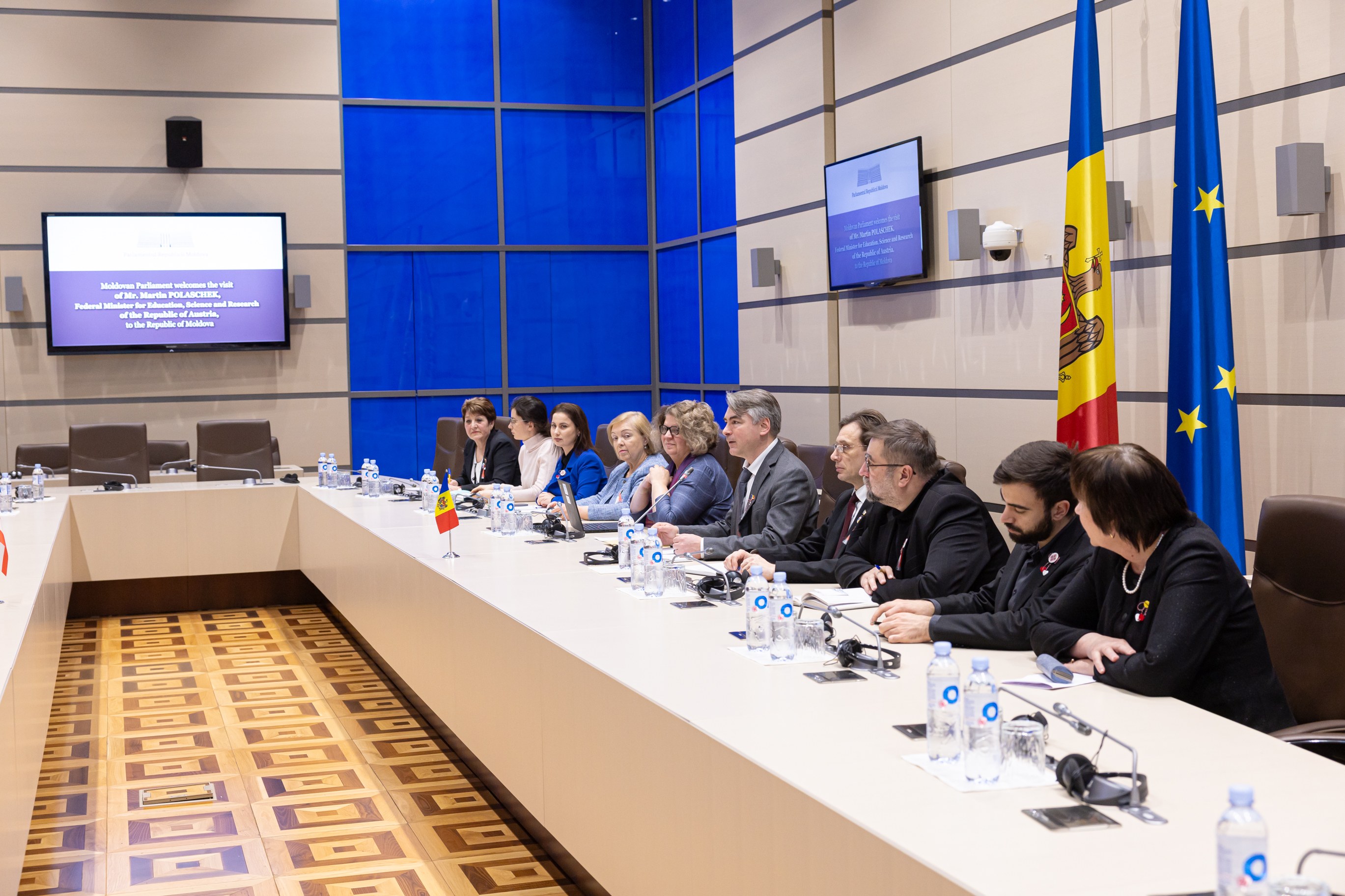 Gruppe von Menschen, die um einen langen Tisch in einer formellen Sitzung mit Flaschen, Gl├Ąsern, Mikrofonen, Papieren und Flaggen im Hintergrund sitzen, ├Ąhnlich einer Sitzung des Europ├Ąischen Parlaments.