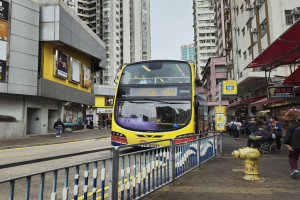 Ein gelber Doppeldeckerbus fährt auf einer von hohen Gebäuden gesäumten Straße, mit Menschen auf dem rechten Gehweg, einem Hydranten und einem Geländer, unter einem klaren blauen Himmel.