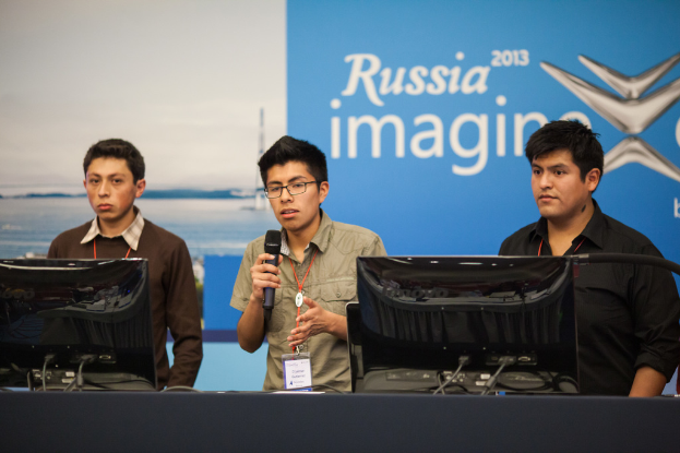 Drei Jungs stehen neben einem Schreibtisch mit zwei Computersystemen, einer spricht in ein Mikrofon und trägt ID-Karten mit roten Tags, vor einer Wand mit einer Werbetafel, auf der 'Russia Imagine 2013' steht.