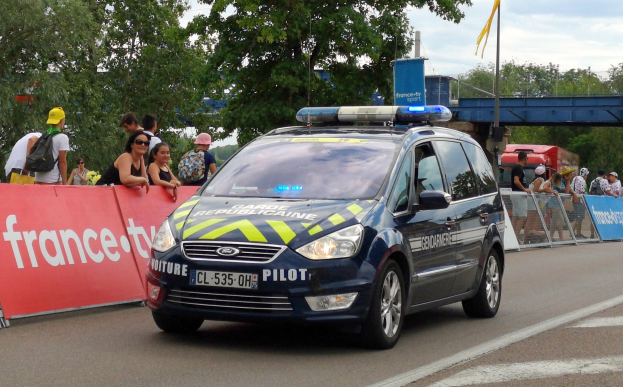 Ein Polizeiwagen fährt auf einer Straße neben einer Menge von Menschen, einige tragen Mötzen und Taschen, mit einem Banner auf der linken Seite, Geländern mit Bannern dahinter und einem Hintergrund aus Bäumen, einer Brücke, einer Fahne auf einem Mast und einem bewölktem Himmel.