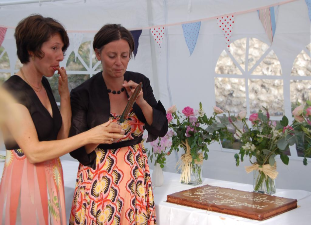 Zwei Frauen stehen neben einem Tisch mit einer Decke, Blumenvasen und einem Kuchen; eine Frau hält ein Messer, mit Flaggen und Fenstern im Hintergrund und einer Wand dahinter.