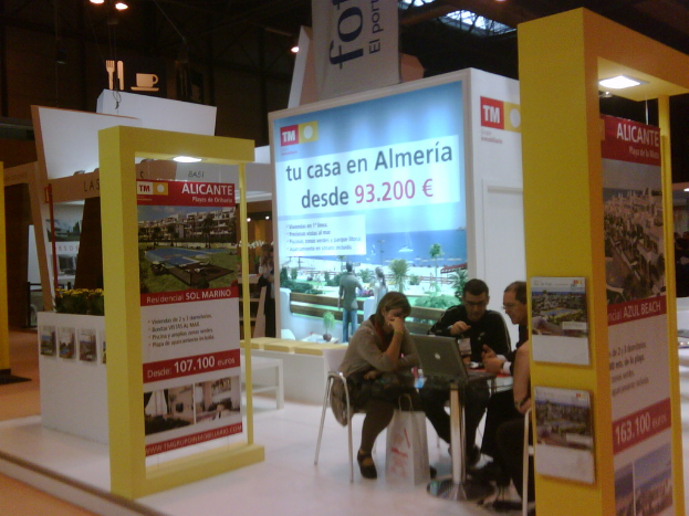 Eine Gruppe von Menschen, die um einen Tisch herum auf Stühlen sitzen und einen Laptop anschauen, mit mehreren Flyern, auf denen verschiedene Werbeanzeigen zu sehen sind.