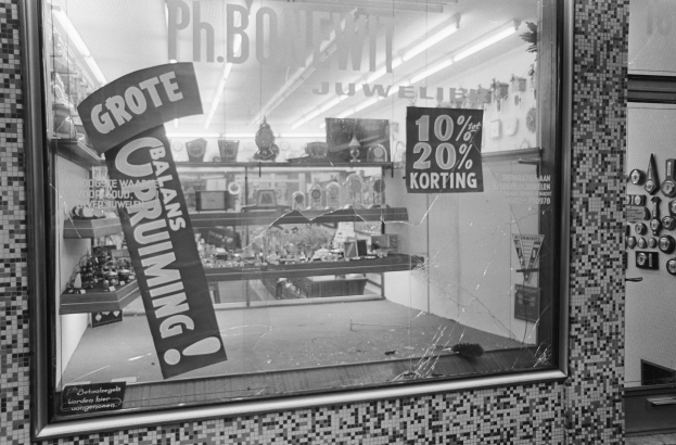 Ein Schwarz-Weiß-Foto eines Ladenschaufensters mit zerbrochener Scheibe, Stickern auf der rechten Seite und einem Gestell mit verschiedenen Gegenständen im Hintergrund.