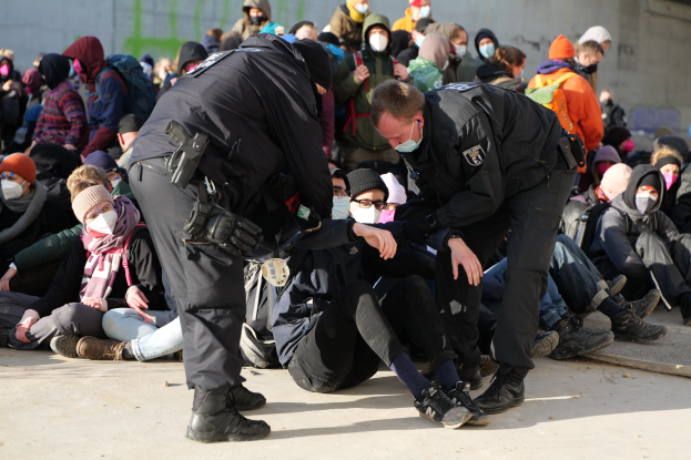 Eine Gruppe von Menschen, einige mit Kappen und Masken, sitzt auf dem Boden mit zwei Polizeibeamten in der Nähe, vor einer Wand mit Schrift, was auf eine Stadtlocation hindeutet.