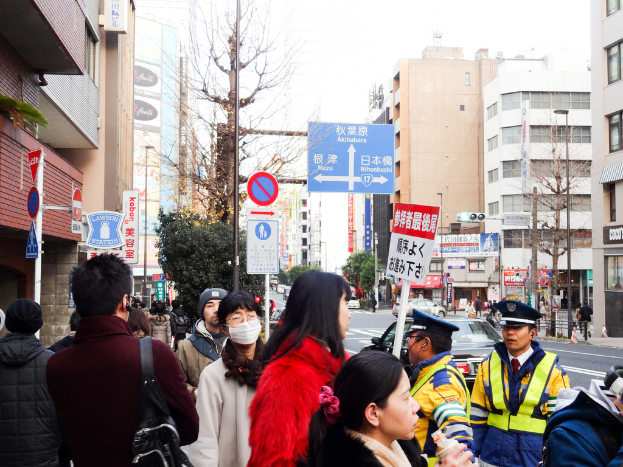 Eine Szene mit vielen Menschen in verschiedenen Kleidern und Mützen, eine Handtasche, eine Haarsträhne, eine Platte, ein Pfahl, Bäume, Gebäude mit Fenstern, eine Straße mit einer weißen Linie und einen weißen Himmel.