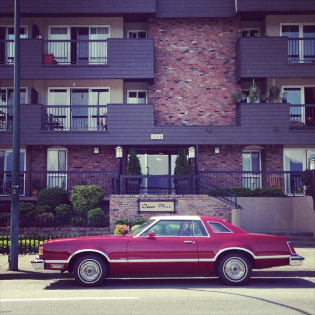Ein rotes Auto steht unten auf dem Bild, mit einem Gebäude in der Mitte, Pflanzen davor und einem Pfahl auf der linken Seite.