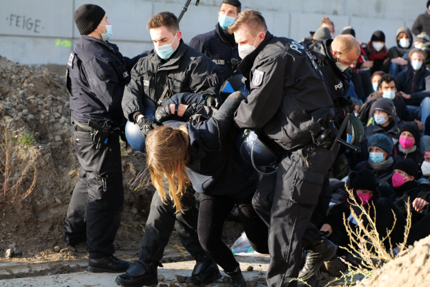 Eine Gruppe maskierter und vermummter Personen, einige bewaffnet, umgibt eine Frau, die von der Polizei eskortiert wird, in einer Stadtkulisse mit Pflanzen, Steinen und einer Wand mit Text, wahrscheinlich protestieren sie gegen Regierungssparmaßnahmen.