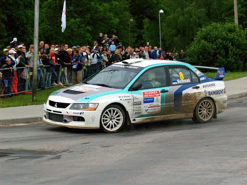 Ein Rallyeauto mit Sponsorenlogos fährt an Zuschauern vorbei, die Kameras halten, auf einer Straße mit Geländern, Polen mit Fahnen und Bäumen im Hintergrund.