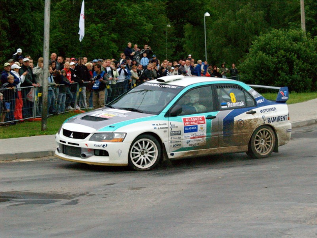 Ein Rallyeauto mit Sponsorenlogos fährt an Zuschauern vorbei, die Kameras halten, auf einer Straße mit Geländern, Polen mit Fahnen und Bäumen im Hintergrund.