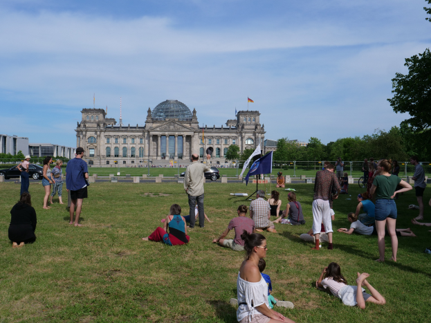 Eine Gruppe von Menschen sitzt auf einem grünen Feld vor dem Reichstaggebäude in Berlin, Deutschland, umgeben von Bäumen, Fahrzeugen und Fahnenstangen.