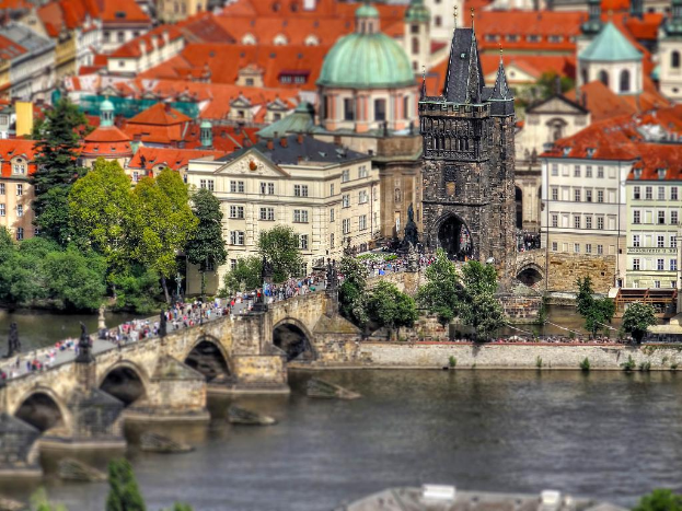 Eine Stadtansicht mit zahlreichen Gebäuden, einem markanten Turm, Bäumen und Menschen, die einen Fluss und eine Brücke überqueren.