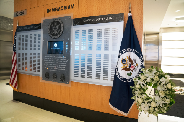Ein Gedenkort in einer Lobby mit Flaggen, einem Kranz und einem Blumenstrauß im Vordergrund sowie einer Wand mit Tafeln und Text, Aufzügen und Deckenleuchten im Hintergrund.