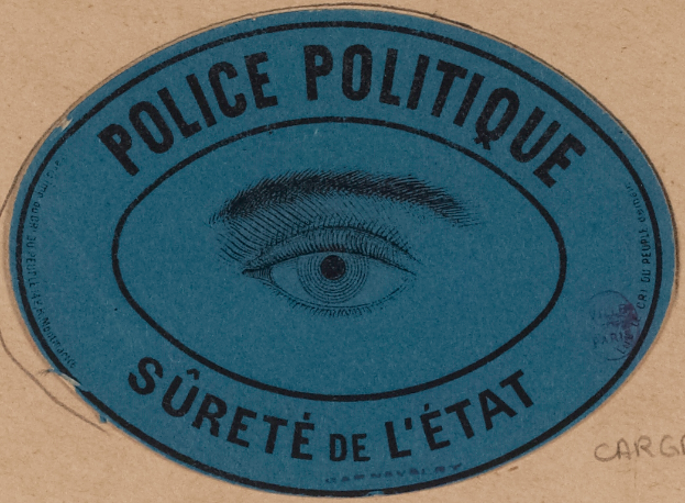 Ein brauner Karton mit einem blauen Aufkleber, der "Polizei Politique Surete de L'État" und ein Auge in der Mitte trägt.