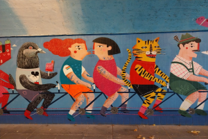 Mural an einer Gebäudeseite, das Menschen beim Radfahren zeigt, mit einer Straße unten und Himmel darüber, Teil einer Londoner Streetart-Serie.