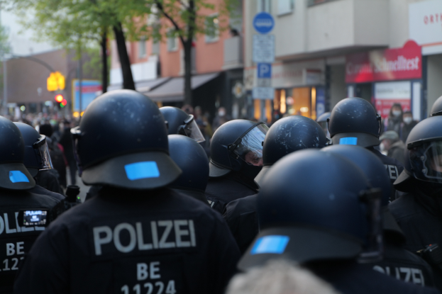 Eine Gruppe von Polizisten in schwarzen Uniformen und Helmen steht vor einer unscharfen Menge und einer Stadtlandschaft mit Gebäuden, Bäumen, Mästen, Verkehrszeichen und Schildern.