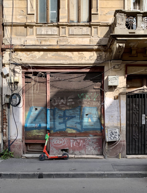 Ein roter Roller neben einem Gebäude mit Fenstern, Rollläden und einem Tor auf einer Straße mit sichtbaren Kabeln und Gegenständen, einschließlich Text unten.