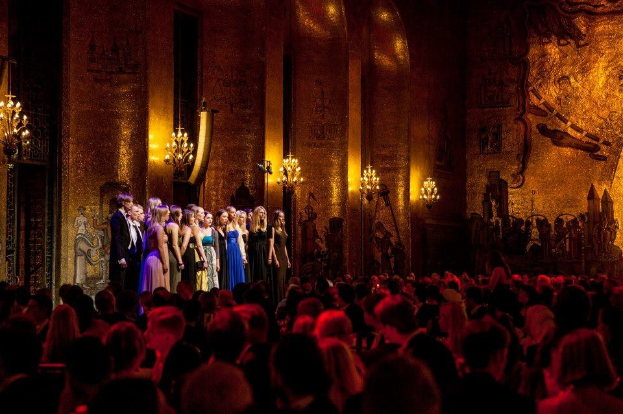 Eine Gruppe von Menschen auf einer hell erleuchteten Bühne im Royal Opera House in London, mit einem Publikum darunter.