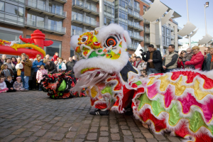 Ein lebendiges chinesisches Neujahrsfest in Amsterdam mit einer Drachenparade und einer Menge Menschen, einige mit Kameras, vor Gebäuden und Laternenmasten unter einem klaren blauen Himmel.