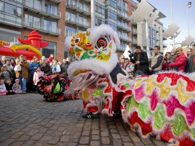 Ein lebendiges chinesisches Neujahrsfest in Amsterdam mit einer Drachenparade und einer Menge Menschen, einige mit Kameras, vor Gebäuden und Laternenmasten unter einem klaren blauen Himmel.