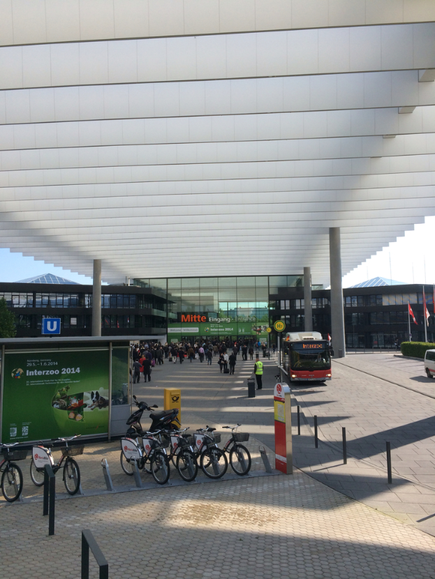 Das Bild zeigt eine Straßenszene mit Fahrrädern und Fahrzeugen auf dem Gehweg, ein Banner auf der linken Seite, Fahrzeuge auf der Straße auf der rechten Seite, Menschen unter einem Dach mit Säulen, Gebäude im Hintergrund und Pfosten mit Flaggen. Es gibt auch Pflanzen auf der rechten Seite des Gehwegs.