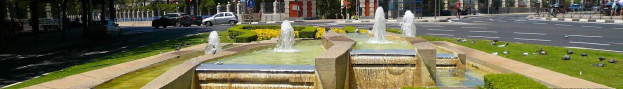 Ein Brunnen auf einer städtischen Straße umgeben von Grünflächen, mit Fahrzeugen und Fußgängern, vor einem klaren blauen Himmel.